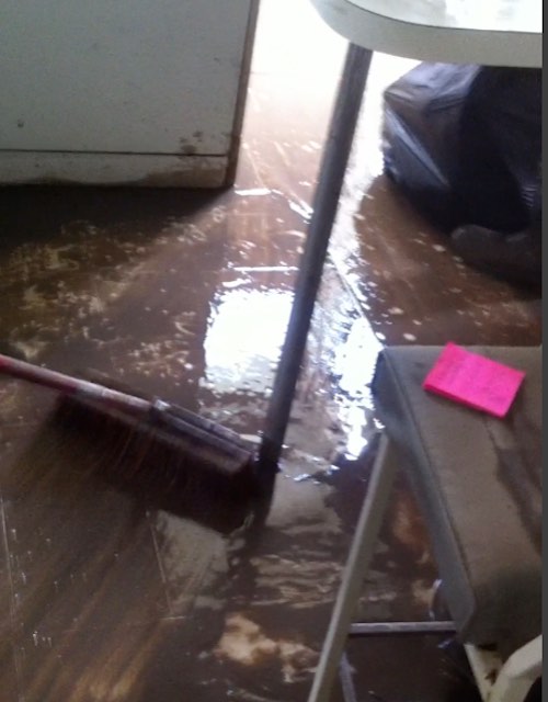 A large broom is sweeping a wet floor covered in dark brown mud. Furniture is scattered about the room. A door is open for the muddy water.
