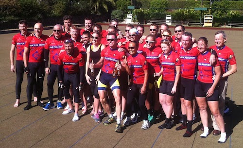 Thirty cyclists standing on a bowling green. They all wear red jerseys with the slogan "Ride for Recovery" on their chests. Everyone is smiling after finishing a 1000km ride.