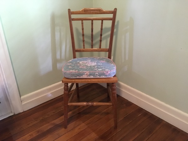 A restored chair sits in the corner of a room. It has a shallow base to sit on. The timber appears newly finished although it is quite old.